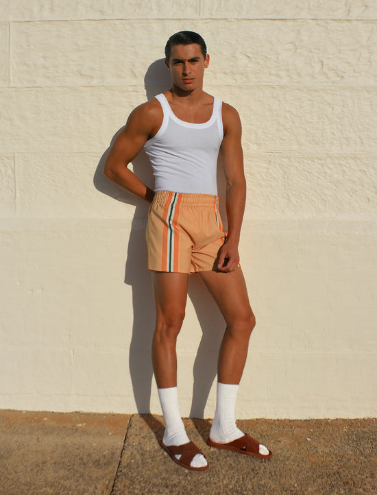 Man wearing a hotel poros white tank top and beige striped summer trunks against a beige wall.
