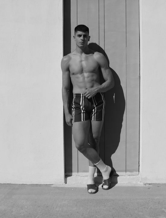 Man standing shirtless wearing striped hotel poros shorts against a white washed wall.