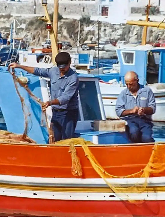 Two greek men on a boat in a harbour with other boats and buildings in the background.