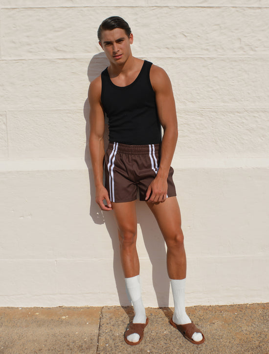 Man wearing a Hotel Poros black tank top and brown shorts with white stripes against a beige wall.