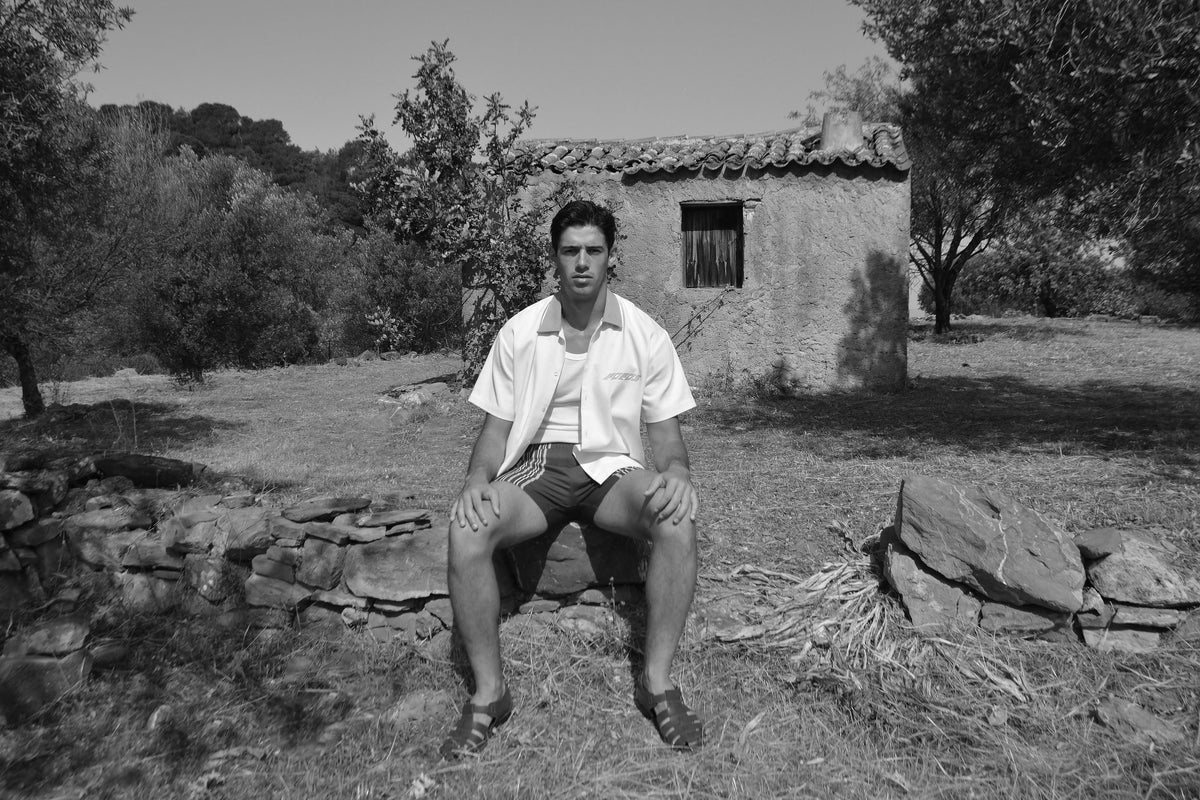 Hotel Poros campaign photograph in black and white of a man sitting outdoors in front of a rustic stone building, wearing the dimitri short-sleeve button-up shirt in white, samos trunk short in navy, and leather sandals.