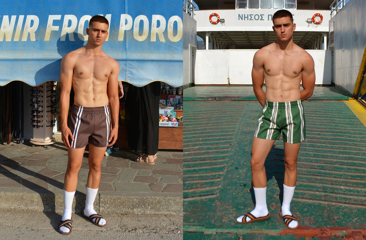 Male model wearing hotel poros swim shorts in green and brown with stripes on the street of Poros island, Greece.
