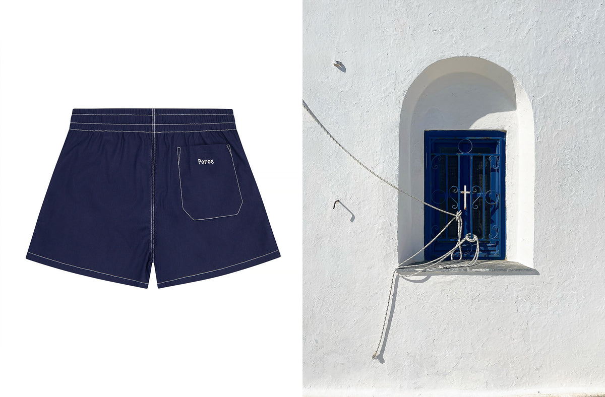 Navy blue linen shorts with white contrast stitching and a small white embroidered poros logo on the back pocket, shown on a white background next to a scenic shot of a white wall with a blue window in greece.
